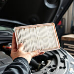 Avotunnelma autotallissa: moottoritilaan kannettava uusi suodatinosa auton huoltoa varten, työkalut ja avoin konepelti taustalla.